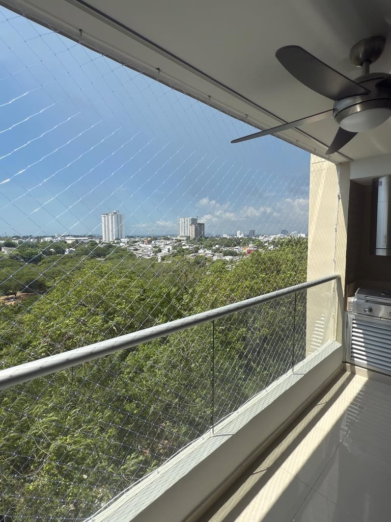 High-rise balcony overlooking green treetops, distant city skyline, and blue sky with ceiling fan above metal railing