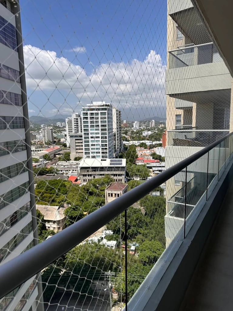 View from high-rise balcony through safety netting overlooking urban cityscape with buildings and blue sky